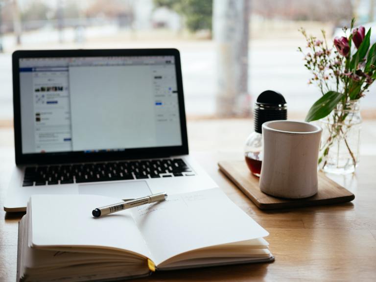 A desk with several items on top. An open laptop, an open notebook with a pen resting on it, a mug and a plant.