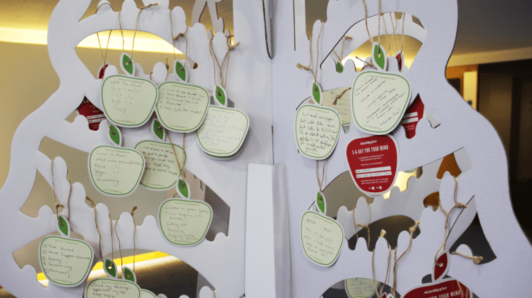 A white cardboard tree, with lots of paper apples hanging from it's branches with string. Each apple has something written on it by hand.