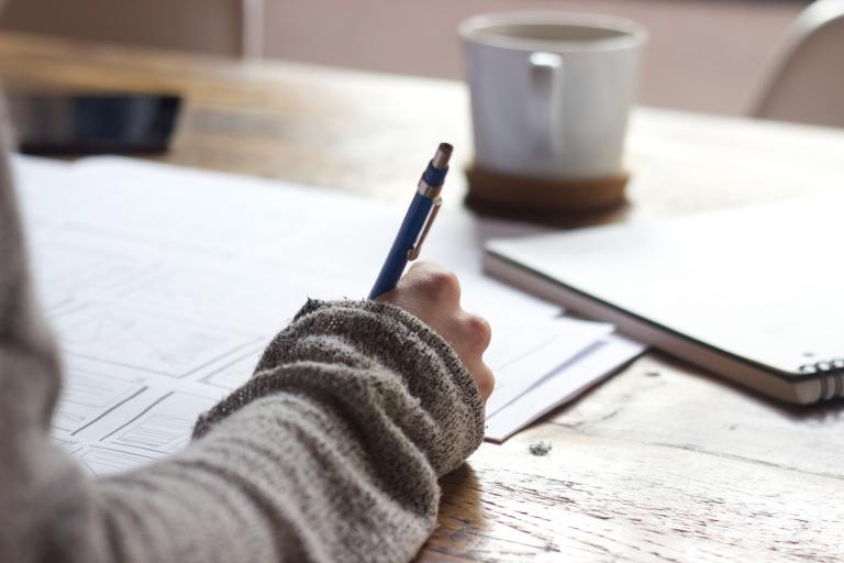 Close up of a person's arm, they are at a desk holding a pen and writing in a notebook. Around the notebook are other pieces of paper and a mug.