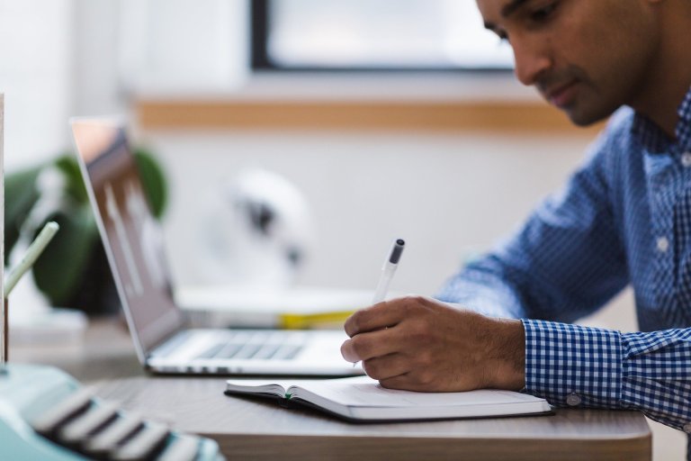 Side view of a man at a desk, holding a pen and writing in a notebook with a laptop next to him.