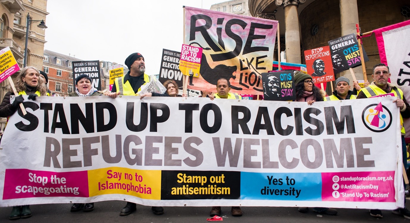 People hold a banner that says 'stand up to racism refugees welcome'. 