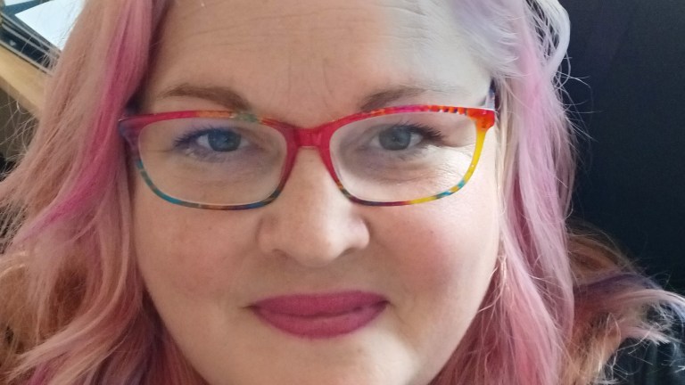 A woman's face, she is wearing colourful glasses and has pink hair, and she is smiling at the camera.