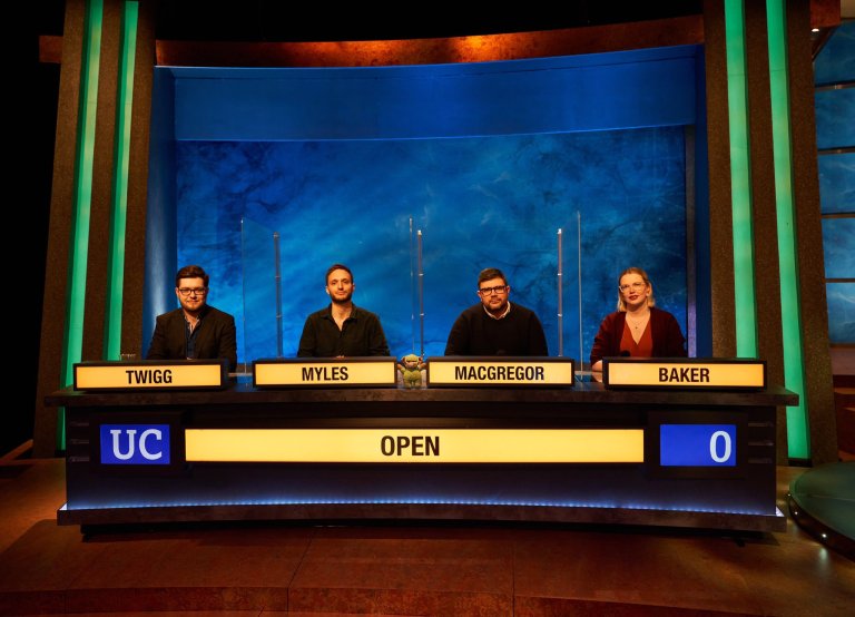 The set of TV show 'University Challenge'. 4 people sat behind their desk as a team with 'Open' lit up under them to signal they are from The Open University'.