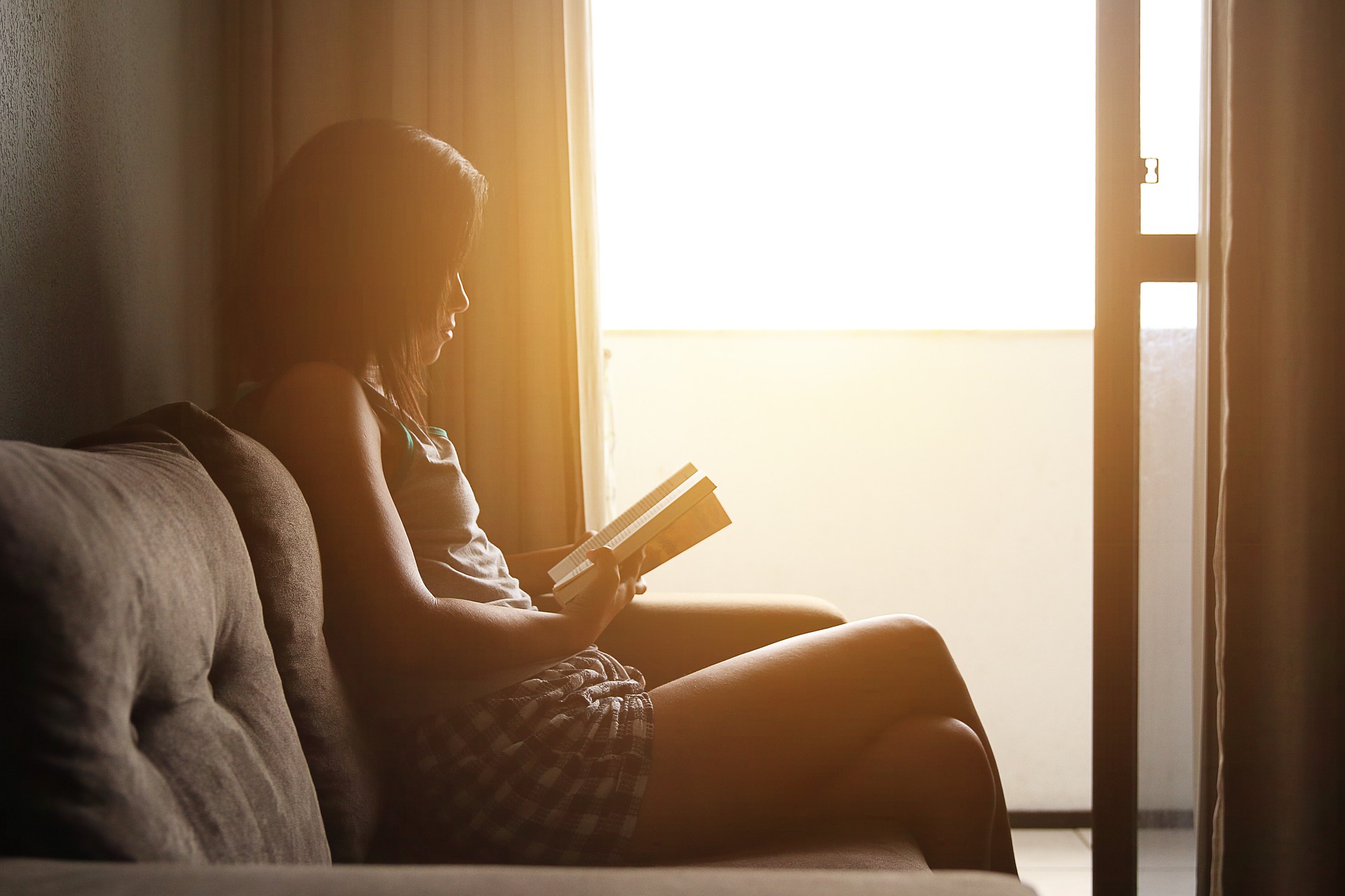 A woman sat by a window on a sofa reading a book with a soft glow from the sun coming through the window,