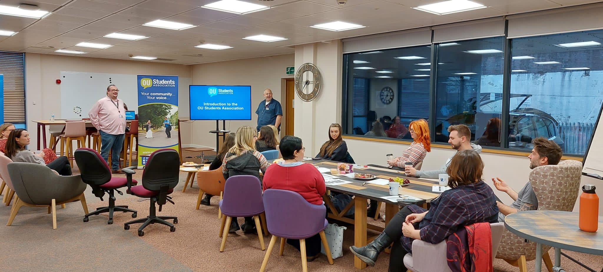 A room full of people in a seminar room sat around tables with a presentation on a screen.