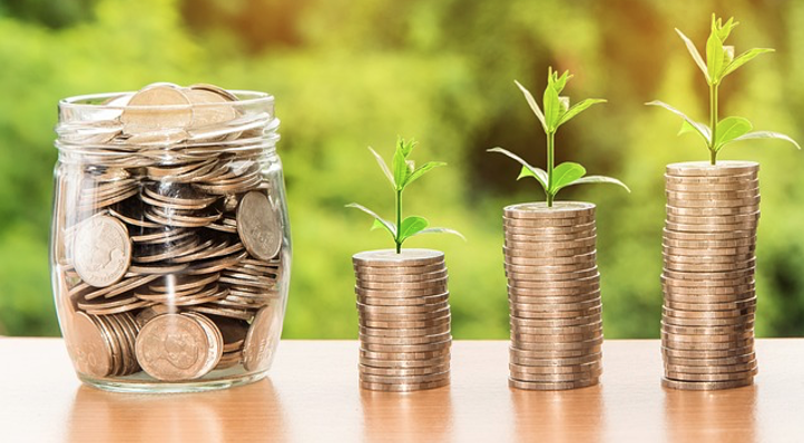A jar full of coins next to 3 small stacks of coins with leaves growing out the top of them.