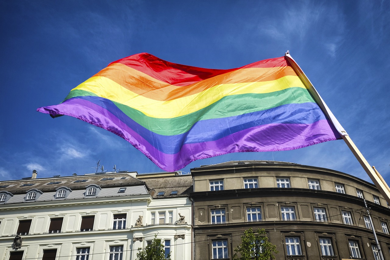 A large rainbow flag, raised on a flag pole, blowing in the wind outside in front of a building.