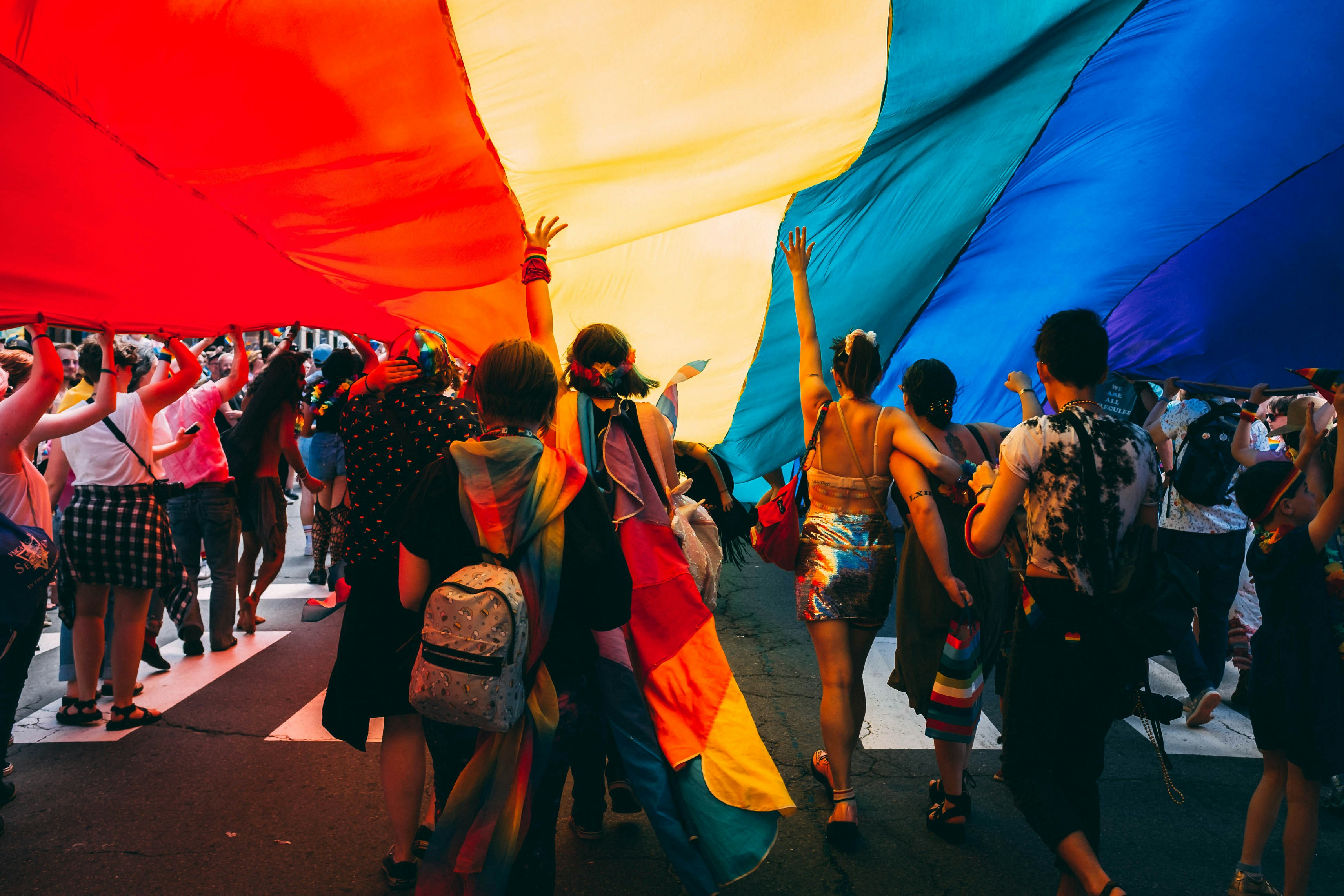 A group of people walking together outside holding a very large rainbow flag above their heads.