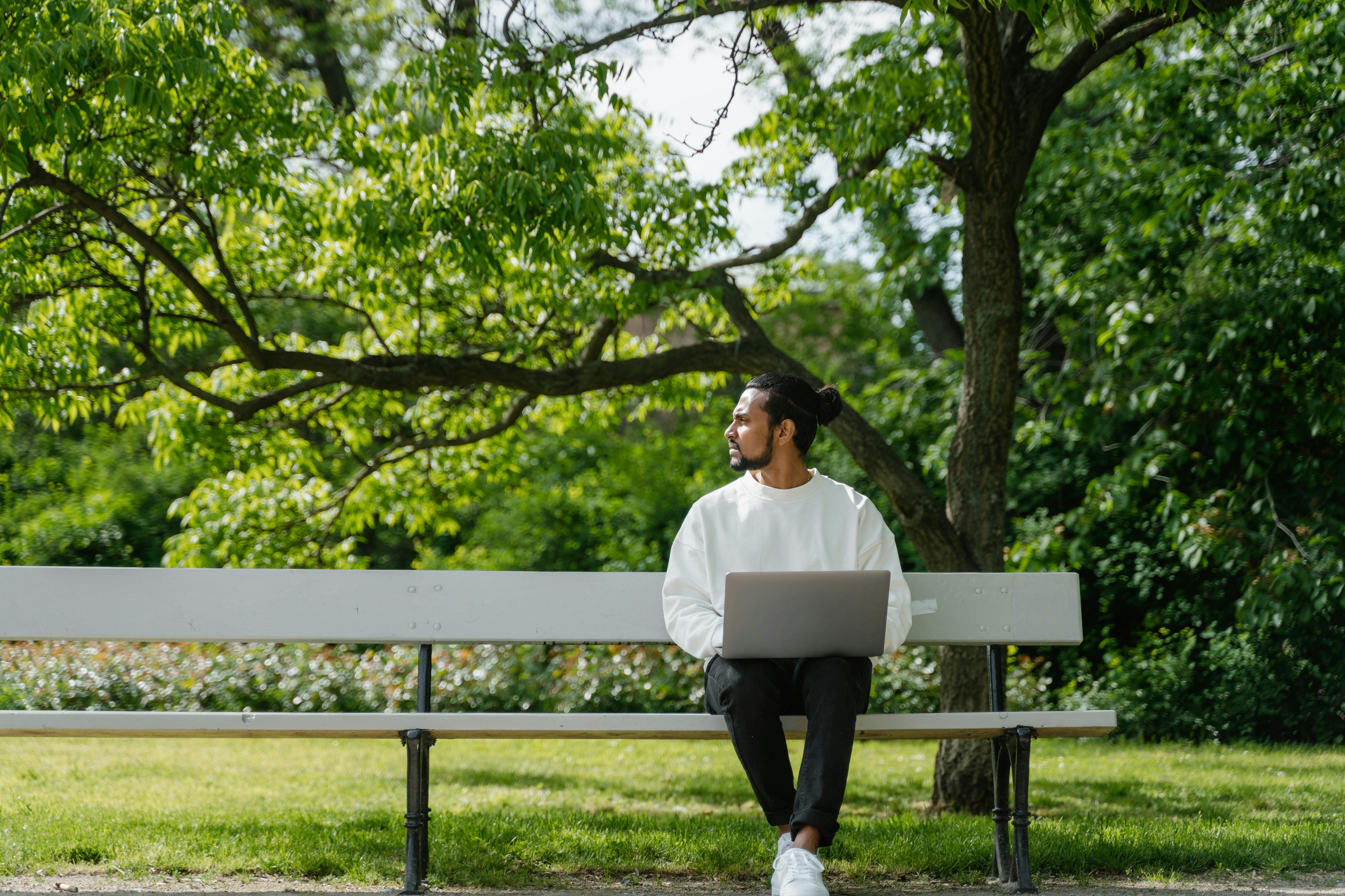 Someone sat on a park bench in the sunshine, working on a laptop and looking into the distance.