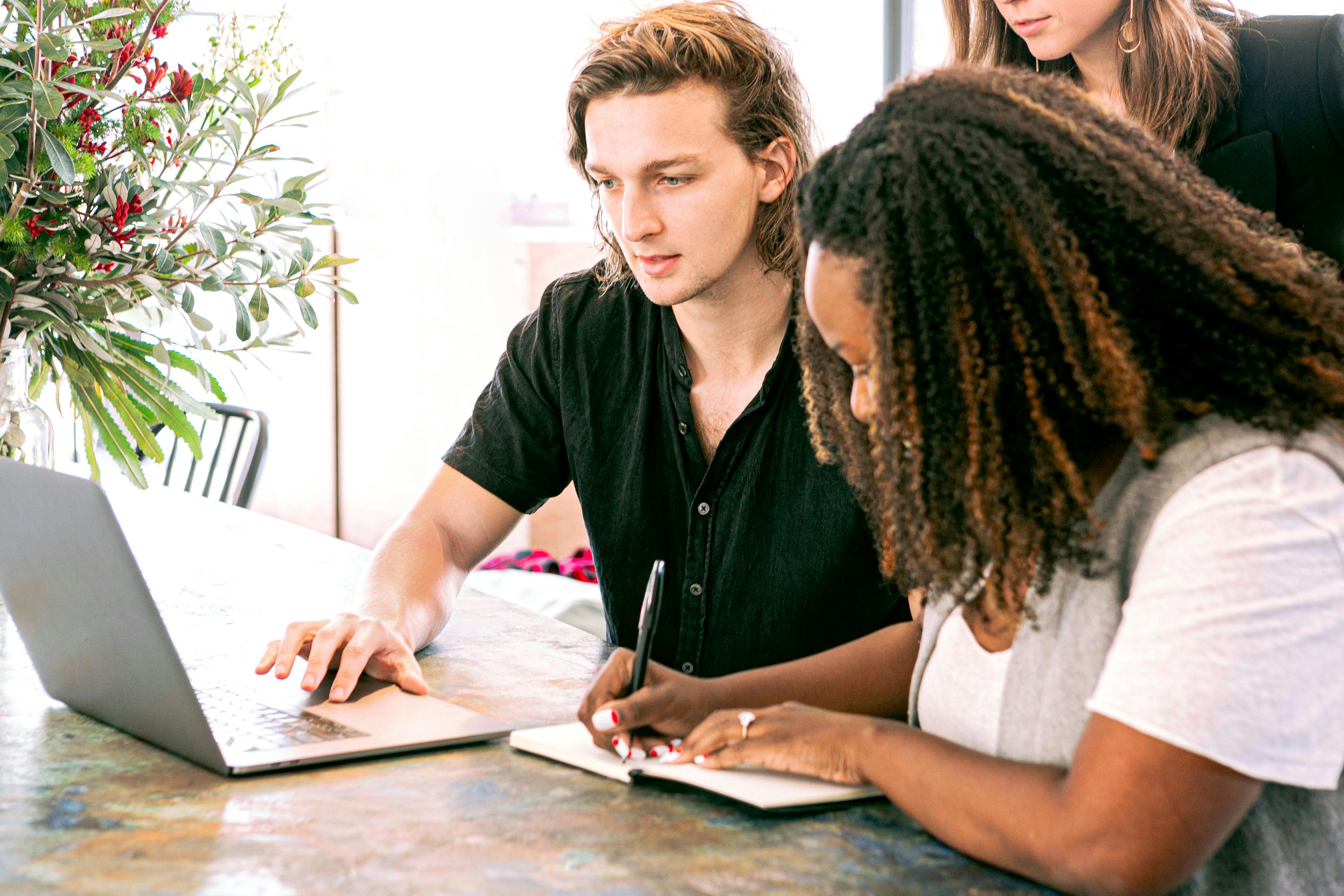 A group of people gathered around a laptop. Someone is working on it and another person is writing in a notebook.
