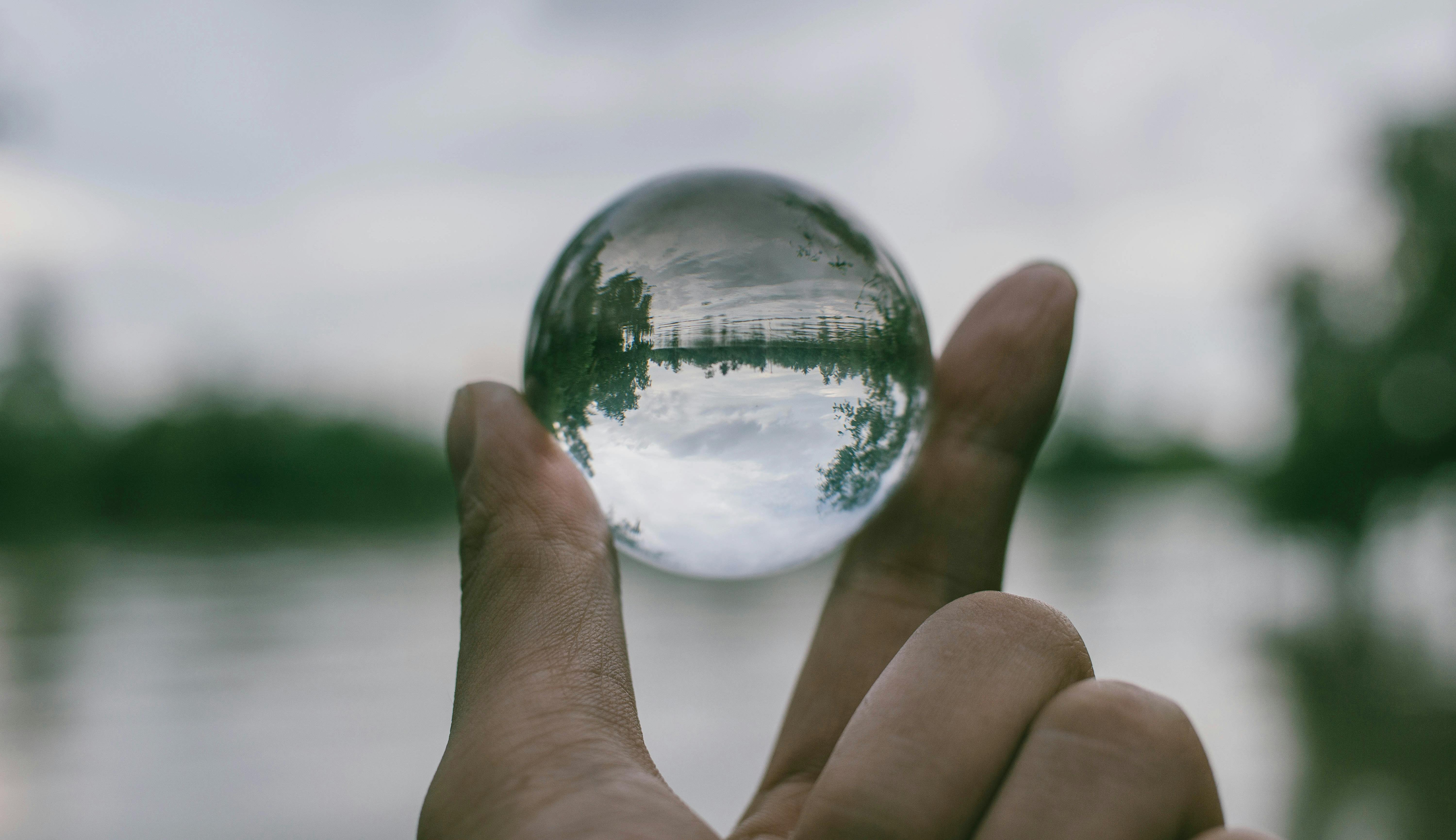 A glass ball held between a thumb and forefinger on a riverside.