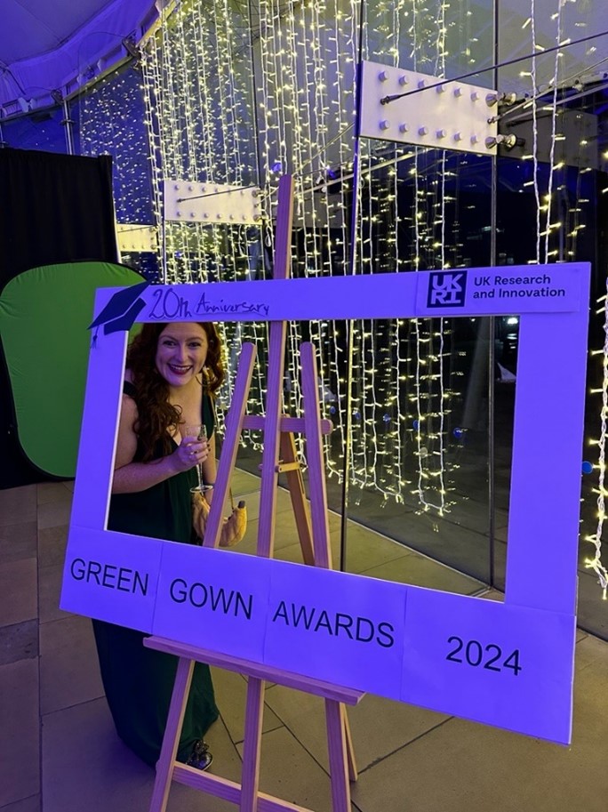 Lou Robinson holding a glass of Prosecco in front of a sparkly curtain. She's standing by a display which says 'Green Gown Awards 2024'.