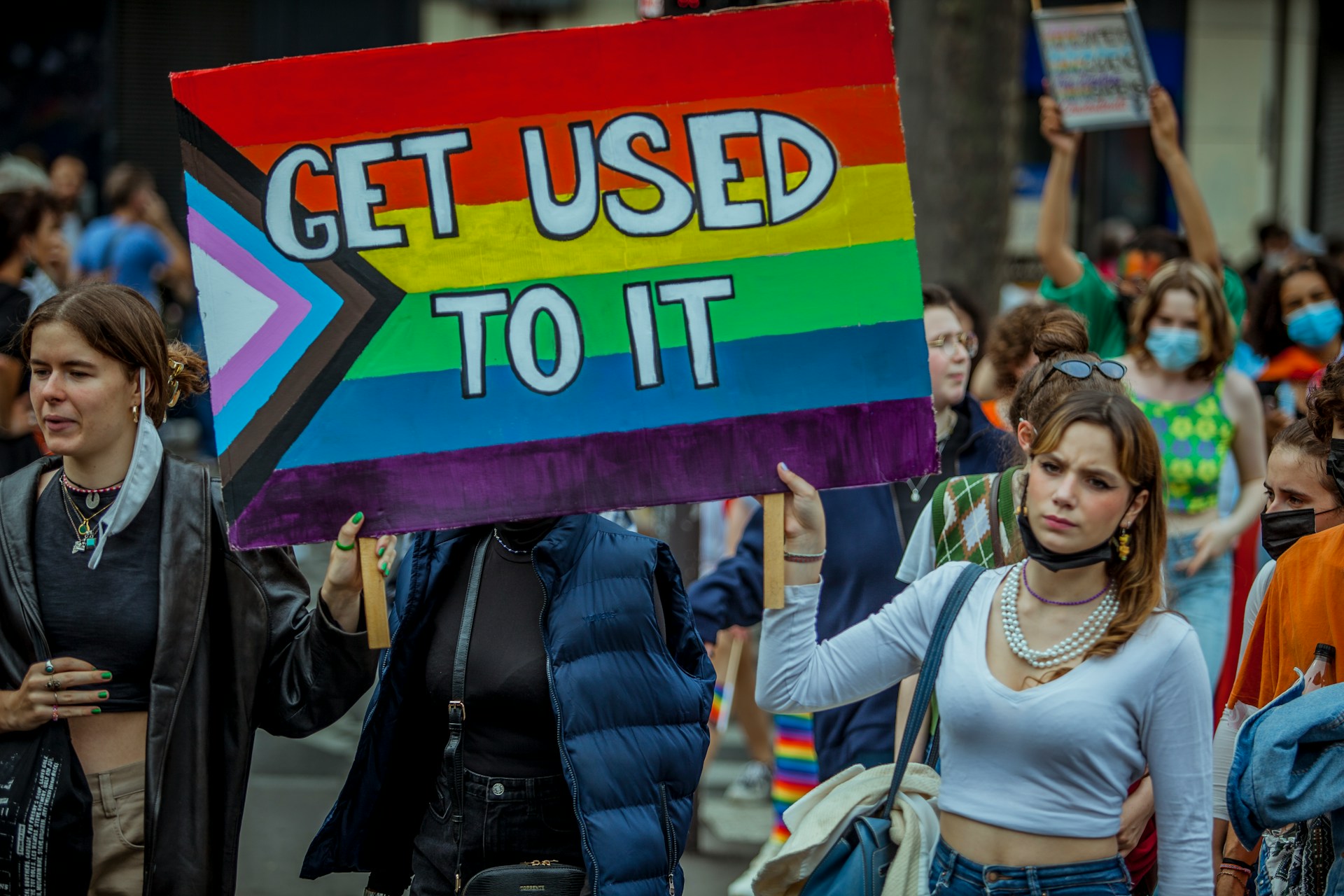 Two people at a rally, holding a LGBT and trans flag, with 'Get used to it' on there.