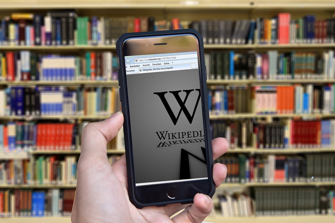 A hand holding up a phone with Wikipedia on it, in front of bookshelves.